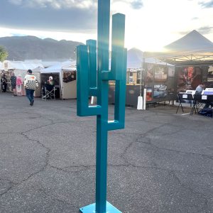 A geometric steel sculpture resembling a simplified cactus, painted bold teal. Made from straight steel piping welded at 90-degree angles, standing tall with clean, angular lines.