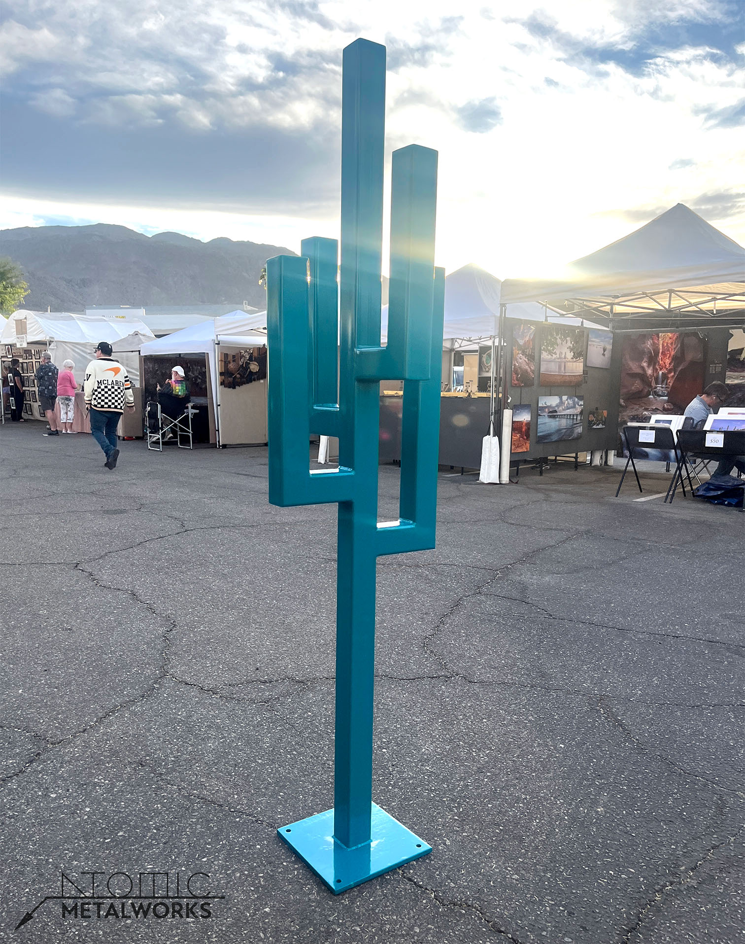 A geometric steel sculpture resembling a simplified cactus, painted bold teal. Made from straight steel piping welded at 90-degree angles, standing tall with clean, angular lines.