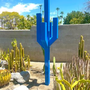 A tall steel sculpture in bright blue, resembling a cactus with arms extending outward at 45 degrees and turning upward at another 45 degrees, creating a modern, angular design.