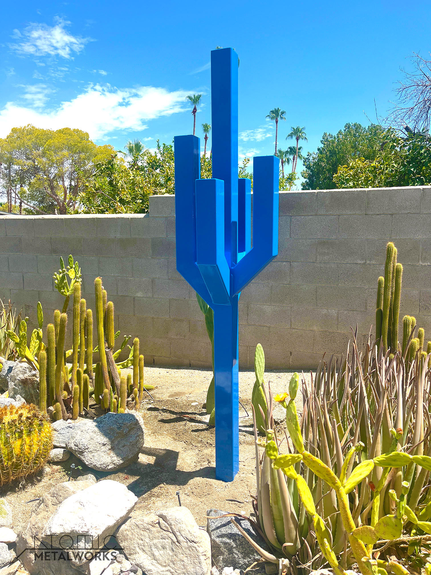 A tall steel sculpture in bright blue, resembling a cactus with arms extending outward at 45 degrees and turning upward at another 45 degrees, creating a modern, angular design.